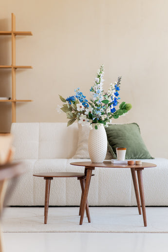 Un salon moderne avec un canapé blanc, un coussin vert et une petite table en bois avec un bouquet de fleurs artificielles en soie Cool Breeze dans un vase blanc, ainsi qu'une tasse à café et des livres, contre un mur beige avec une étagère minimaliste.