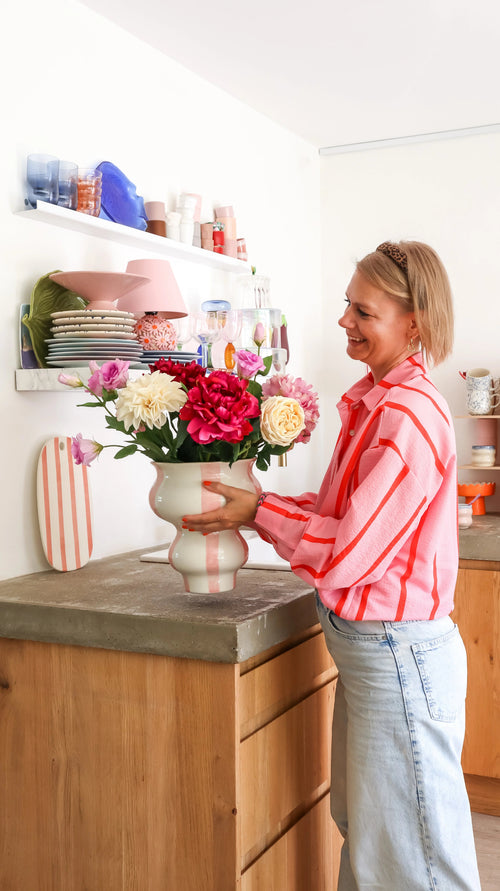 Une femme vêtue d'une chemise à rayures roses dispose le luxueux cadeau floral, un bouquet de fleurs artificielles en soie Dream Delight, dans un vase rayé sur le comptoir, avec des étagères d'assiettes et de verres visibles derrière elle.