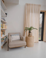 Un coin lecture cosy avec un fauteuil beige, une table d'appoint ronde en osier sur laquelle est posée la plante artificielle Baby Cycas Palm 60 cm, des bibliothèques et des voilages qui pendent au-dessus d'une porte vitrée qui s'ouvre sur le jardin.