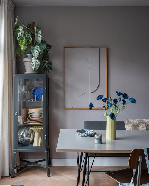 La salle à manger moderne comprend une vitrine en verre gris dans laquelle sont exposées de la vaisselle et des plantes, des décorations murales minimalistes et une table sur laquelle se trouve la fleur artificielle Coquelicot 73 cm bleu foncé dans un vase jaune. L'espace est complété par un mélange éclectique de chaises, le tout sur fond de murs gris et de parquet.