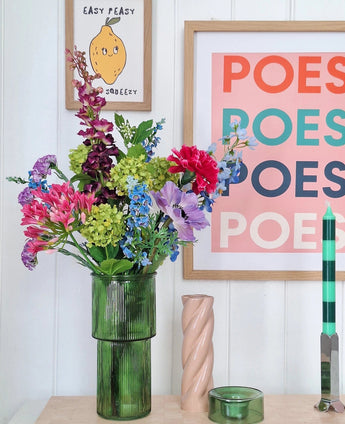 Un vase en verre vert contient un bouquet de fleurs artificielles en soie Blossom Breeze sur la table. Derrière, sont accrochées des images encadrées – une avec un citron et « EASY PEASY », une autre avec « POESY » – ajoutant un charme supplémentaire. A proximité se trouvent une bougie verte et une bougie spirale rose de Blossom Breeze.