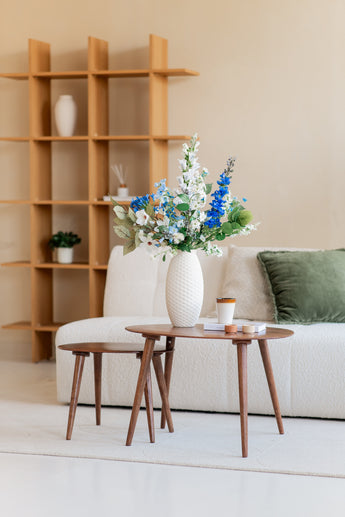 Un salon moderne avec un canapé structuré blanc, un coussin vert, des tables basses en bois et un vase blanc avec le bouquet de fleurs artificielles en soie Cool Breeze. Une planche de bois à la décoration minimaliste complète cet environnement serein.