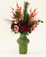 Le bouquet de fleurs artificielles Silk Cosy Winter présente un vase vert nervuré avec des fleurs rouge foncé et roses, des baies orange, des branches de pin et des feuilles vertes - parfait comme décoration automne-hiver sur un fond clair.