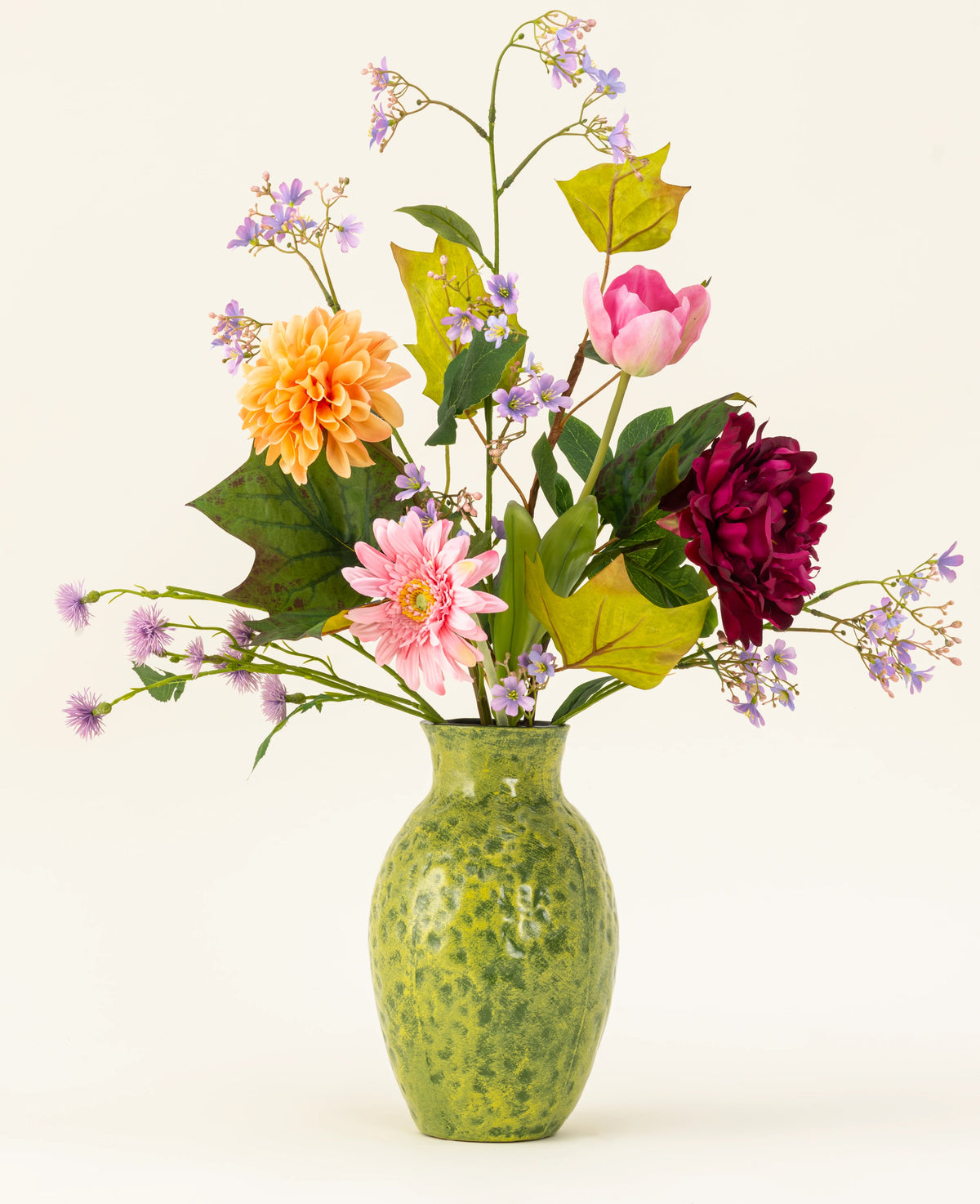 Un vase vert moucheté montre le bouquet de fleurs artificielles en soie Love in Bloom, avec des fleurs roses, orange et rouge foncé avec un feuillage violet et vert sur un fond clair.