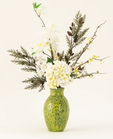 Le bouquet de fleurs artificielles en soie Earthlight est disposé dans un vase texturé vert, avec des hortensias blancs, de délicates fleurs blanches et un feuillage vert-brun sur un fond clair uni.