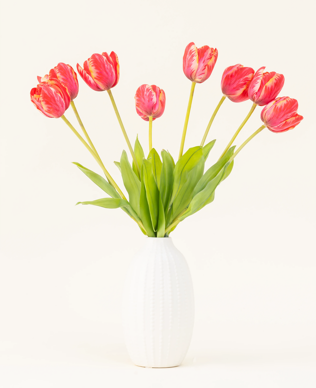 Le bouquet de fleurs artificielles en soie French Tulip rouge est disposé dans un vase en céramique blanche, avec huit tulipes rouges et jaunes aux feuilles vertes sur un fond uni clair.