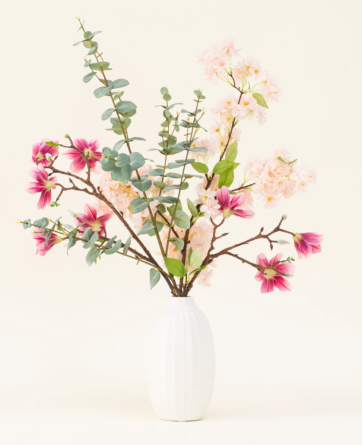 Dans un vase à structure blanche se trouve le bouquet de fleurs artificielles en soie Featherlight Florals avec des fleurs en soie rose tendre et magenta foncé, ainsi que des feuilles d'eucalyptus vertes sur de longues tiges, sur un fond clair.