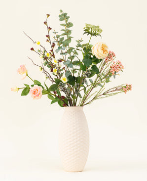 Un vase blanc texturé présente le bouquet de fleurs artificielles en soie Blush Blossom, avec de vraies fleurs – roses rose pâle, fleurs jaunes et feuillage vert – sur un fond clair uni.