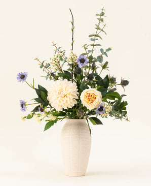 Un vase en céramique blanche contient le bouquet de fleurs artificielles en soie Petal Poetry, avec des dahlias et des roses de couleur crème, des fleurs violet clair, des chardons verts et toutes sortes de verdure sur un fond clair uni.