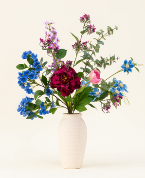 Un vase blanc texturé présente le bouquet de fleurs artificielles en soie Painted Petals, avec des fleurs en soie bleues, violettes, roses et rouges vibrantes avec des feuilles vertes et des branches d'eucalyptus sur un fond clair uni.