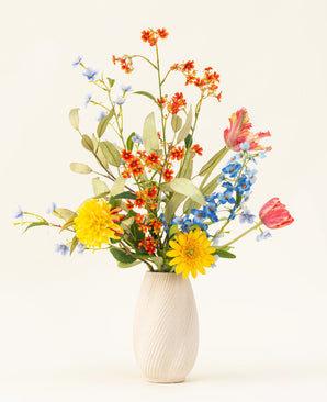 Le bouquet de fleurs artificielles en soie Lemonade Garden comprend des tournesols teintés, des tulipes roses, des delphiniums bleus, des fleurs d'oranger et du feuillage vert dans un vase beige sur un fond clair uni.