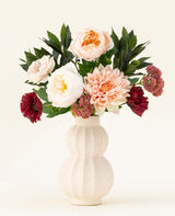 Un vase en céramique de couleur crème avec structure montre le bouquet de fleurs artificielles en soie Jewel Bloom avec des fleurs Real Touch roses, blanches et rouge foncé et des feuilles vertes sur un fond clair uni.