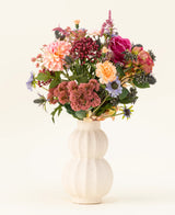 Dans un vase en céramique blanc incurvé se trouve le bouquet de fleurs artificielles en soie Pretty Pastel - un mélange coloré de fleurs durables telles que des roses roses, des dahlias, des fleurs violettes et oranges et de la verdure - magnifiquement disposées sur un fond clair.