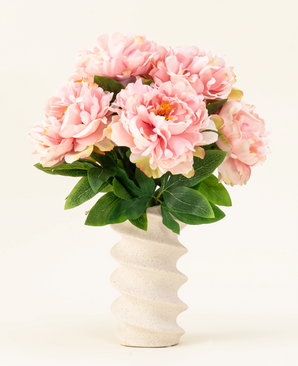 Le bouquet de fleurs artificielles en soie Peony Soft Pink présente de grandes pivoines rose tendre et des feuilles vertes dans un vase en céramique ondulé de couleur crème, magnifiquement mis en valeur sur un fond clair uni.