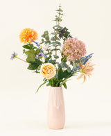 Le bouquet de fleurs artificielles en soie Earth & Petal est un vase rose pastel avec des fleurs en soie réalistes en orange, rose et jaune, ainsi que des accents verts et violets, représentés sur un fond clair.