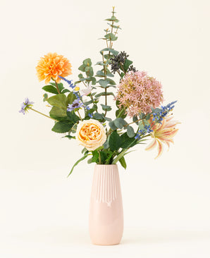Le bouquet de fleurs artificielles en soie Earth & Petal est un vase rose pastel avec des fleurs en soie réalistes en orange, rose et jaune, ainsi que des accents verts et violets, représentés sur un fond clair.
