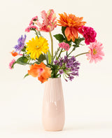 Un vase en céramique rose pâle contient le bouquet de fleurs artificielles en soie Candy Petals avec des fleurs en soie orange vif, jaune, rose et violet sur un fond blanc cassé uni.