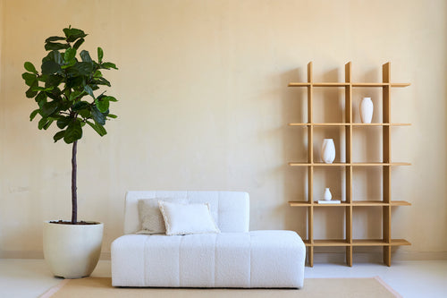 Salon minimaliste avec un canapé blanc, deux coussins et une plante artificielle Ficus Tobacco Plant Deluxe 180 cm à côté d'une étagère ouverte en bois avec des vases blancs, le tout contre un mur beige et sur un tapis clair.