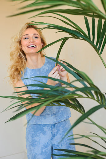 Une femme souriante aux longs cheveux blonds, vêtue d'un haut sans manches bleu clair et d'un pantalon assorti, se tient derrière la plante artificielle Kentia Palm 180 cm et touche de manière ludique l'une des feuilles vertes.