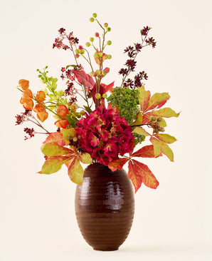 Dans un vase marron nervuré se trouve le bouquet « Fleurs artificielles en soie Ambre chaud », avec des fleurs et des feuilles rouges, oranges et vertes vibrantes sur de longues tiges, sur un fond clair uni.