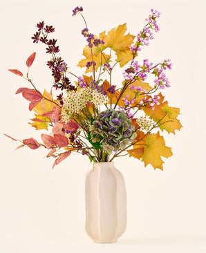 Het boeket ‘Wild Amethyst’ van Zijden kunstbloemen, met herfstbladeren, paarse en witte bloemen en groene hortensia’s, staat in een witte keramische vaas tegen een effen, lichte achtergrond.