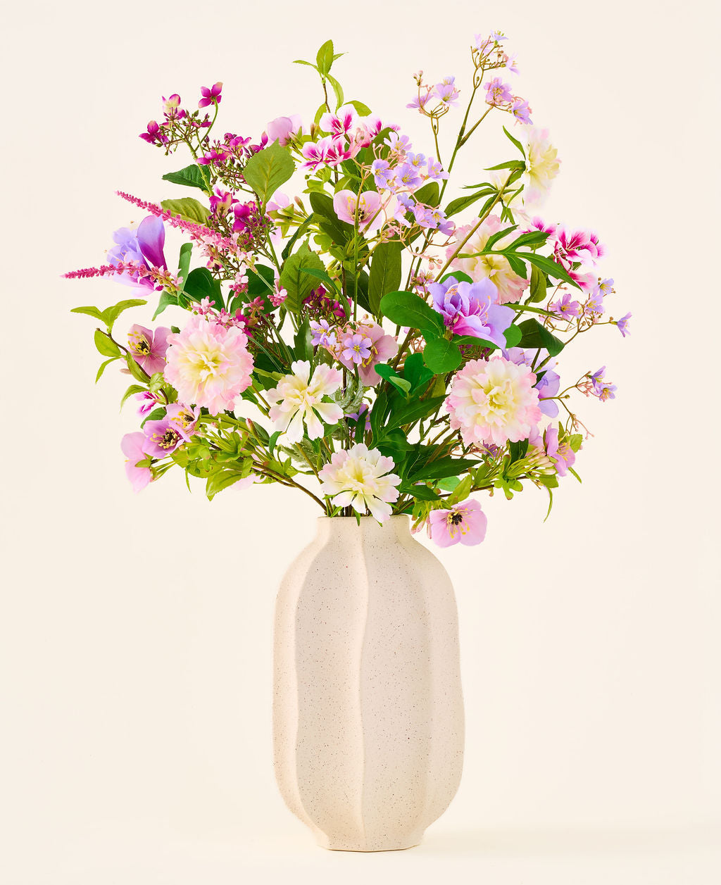Un vase en céramique blanche contient un bouquet coloré de fleurs roses, violettes et blanches de la collection « Bouquet de fleurs artificielles en soie Royal Bloom », rehaussé de feuilles vertes et présenté sur un fond uni et clair.