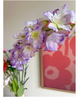 Gros plan de la fleur artificielle Delphinium 105 cm violette, disposée dans un vase, avec un tableau floral rose et rouge sur le mur en fond.