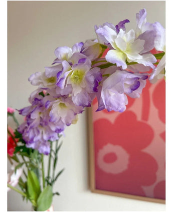 Gros plan de la fleur artificielle Delphinium 105 cm violette, disposée dans un vase, avec un tableau floral rose et rouge sur le mur en fond.