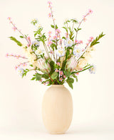Dans un vase à structure beige se trouve le bouquet de fleurs artificielles en soie Lots of Love XL, un bouquet noué à la main de fleurs en soie roses, blanches et bleues avec des feuilles vertes et de longues tiges sur un fond clair.
