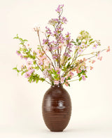 Un vase en céramique marron montre le bouquet de fleurs artificielles en soie Lilas, un bouquet de soie grand et élégant avec des fleurs roses, violettes et lilas avec des feuilles vertes et de fines branches sur un fond clair uni.