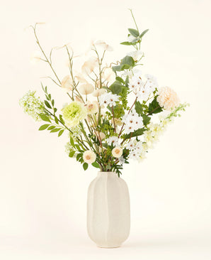 Le bouquet de fleurs artificielles en soie Claire de Lune présente de délicates fleurs blanches, jaune pâle et vertes dans un vase en céramique blanche, accentuées par une verdure luxuriante sur un fond clair.