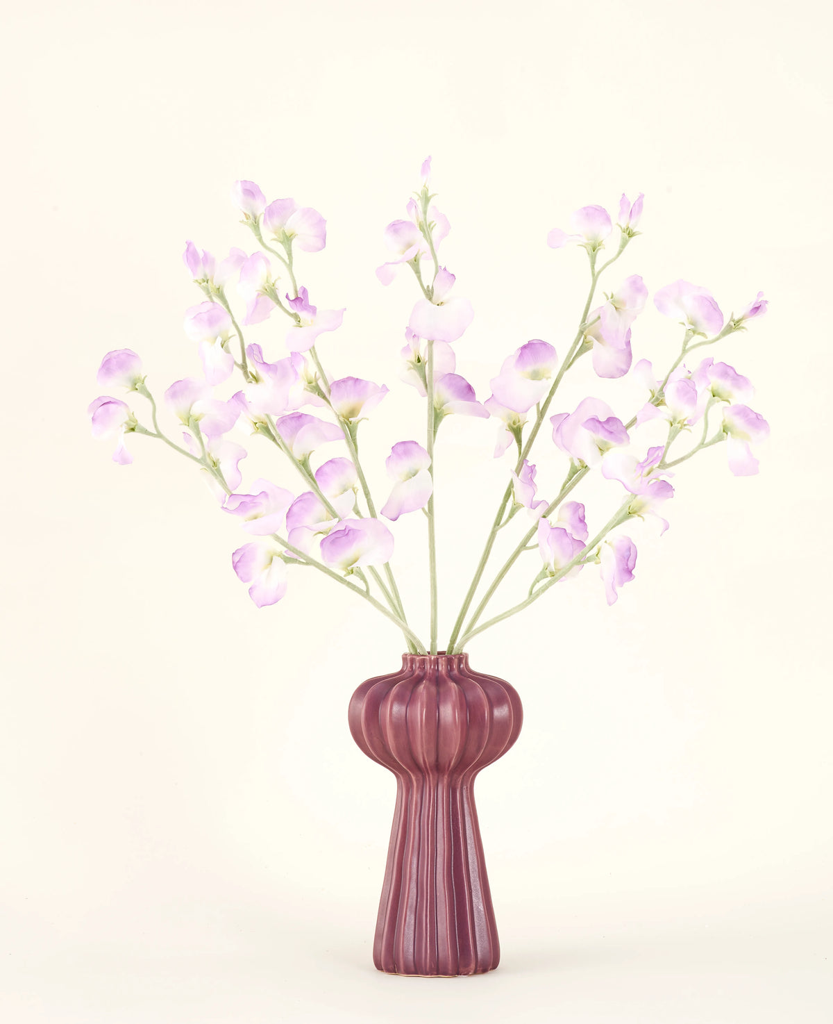 Le bouquet de fleurs artificielles en soie Pois lilas présente de longues tiges de fleurs délicates violet clair et de feuilles vertes dans un vase en céramique mauve aux stries verticales, sur un fond uni blanc cassé.