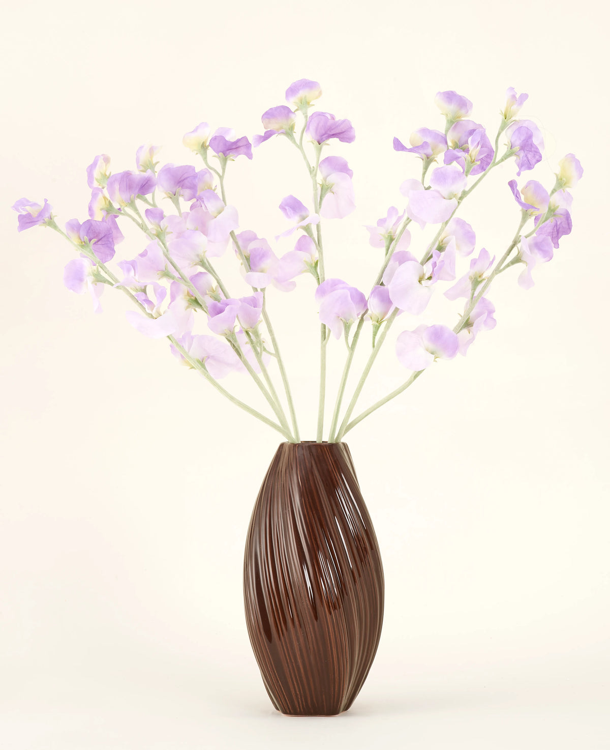 Un vase marron texturé contient le bouquet de fleurs artificielles en soie, des fleurs en soie violet-violet clair avec des tiges verdâtres sur un fond uni et clair.