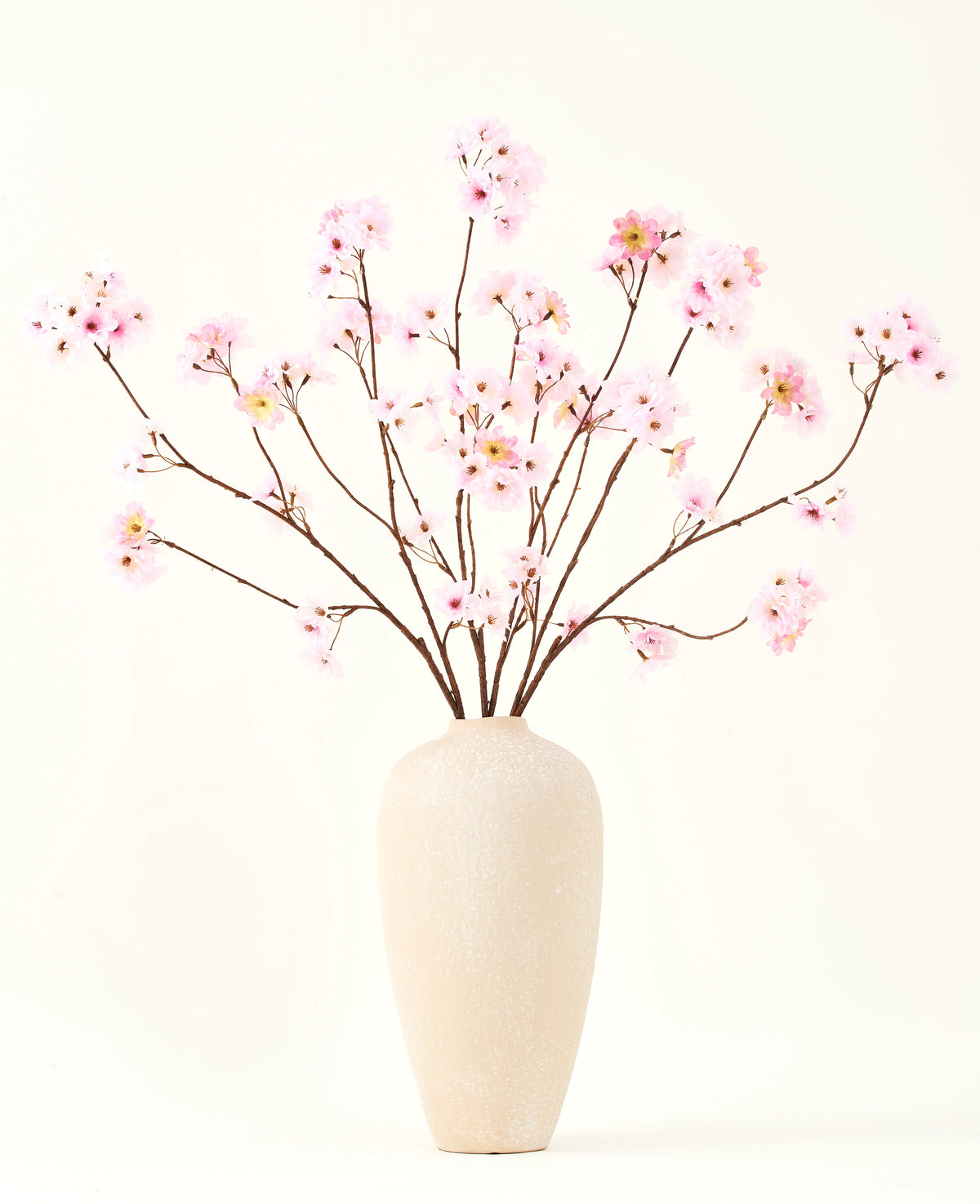 Le bouquet de fleurs artificielles en soie Fleur de cerisier rose 114 cm a des branches longues et fines avec des bouquets de fleurs rose clair dans un vase en céramique beige, qui forment un contraste saisissant sur le fond blanc uni.