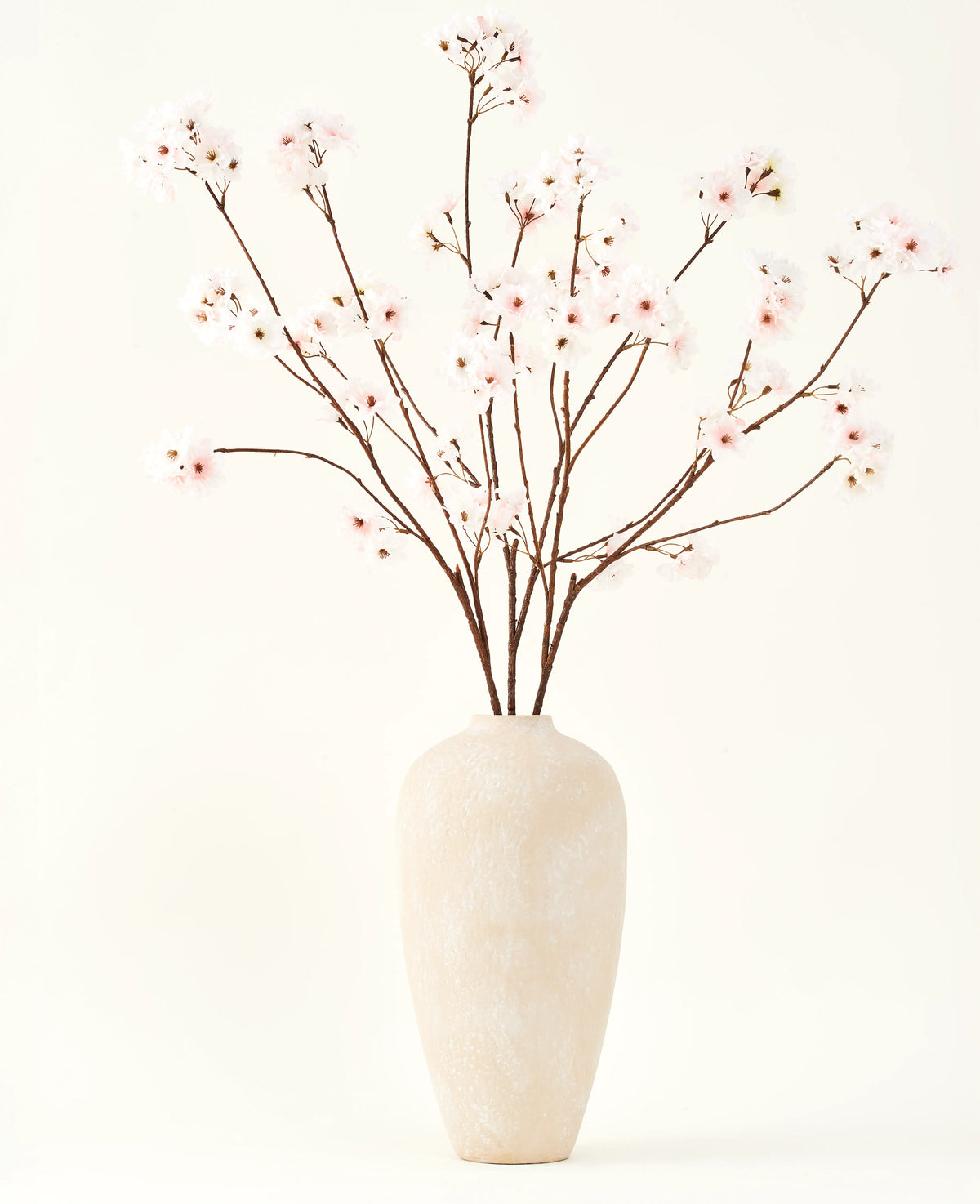 Le bouquet de fleurs artificielles en soie Cherry Blossom Blush 114 cm présente des branches hautes et délicates dans un vase beige sobre, qui apportera une touche douce et élégante à votre intérieur sur un fond uni et clair.