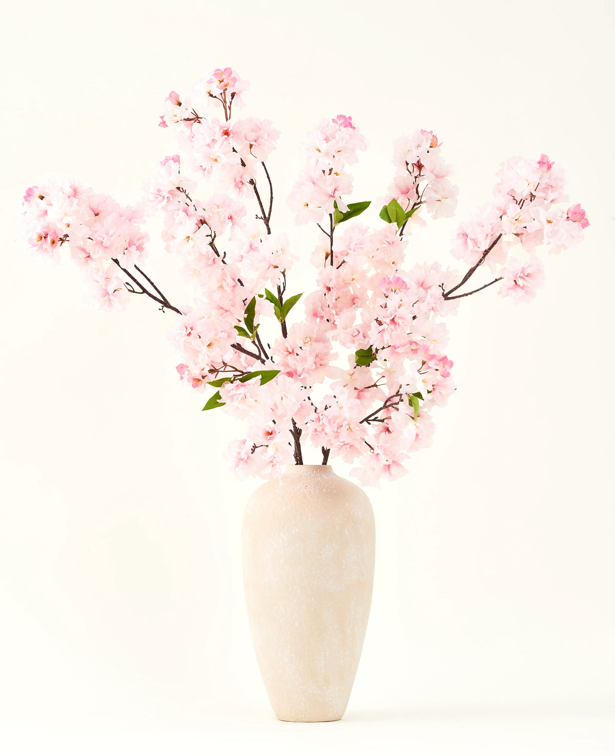 Le bouquet de fleurs artificielles en soie Fleur de cerisier rose 119 cm, disposé dans un vase en céramique beige aux délicates feuilles vertes, crée un accent intérieur serein sur un fond clair simple.