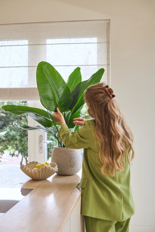 Une femme aux longs cheveux ondulés, vêtue d'un costume vert, s'occupe d'une plante artificielle Strelitzia Real Touch Deluxe de 90 cm posée sur un comptoir à côté d'un bol de citrons dans une pièce lumineuse avec une fenêtre.