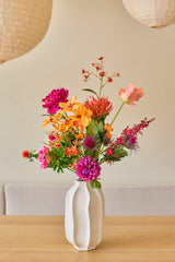 Dans un vase en céramique blanche se trouve le bouquet de fleurs artificielles en soie 'Magnus di Lambo', un arrangement coloré de fleurs artificielles roses, orange et jaunes avec des feuilles vertes, posé sur une table en bois sur un fond beige.