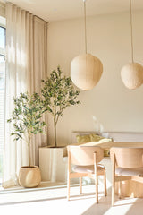 Une salle à manger lumineuse et minimaliste est meublée de deux chaises en bois clair, d'une petite table, de lanternes en papier et de l'olivier artificiel de 180 cm, qui ajoute une touche de style méditerranéen. Des arbres d'intérieur en pot, des rideaux beiges translucides et la lumière du soleil pénétrant à travers les grandes fenêtres complètent cet espace de détente.