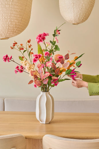Quelqu'un avec une manche verte dispose le bouquet « Fleurs artificielles en soie pêche sauvage », avec des fleurs d'été colorées, dans un vase blanc sur une table en bois clair, avec des lanternes en papier et un canapé beige en arrière-plan.