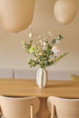 Sur une table en bois se trouve un vase blanc avec un élégant bouquet de fleurs artificielles de Zijden, appelé Claire de Lune. Il y a deux chaises beiges et des lanternes en papier suspendues au-dessus de la table. Le fond se compose d'un mur crème doux avec un canapé avec des coussins, créant une atmosphère minimaliste et cosy rappelant l'atmosphère sereine de Claire de Lune.