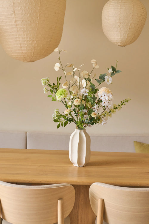 Sur une table en bois se trouve un vase blanc avec un élégant bouquet de fleurs artificielles de Zijden, appelé Claire de Lune. Il y a deux chaises beiges et des lanternes en papier suspendues au-dessus de la table. Le fond se compose d'un mur crème doux avec un canapé avec des coussins, créant une atmosphère minimaliste et cosy rappelant l'atmosphère sereine de Claire de Lune.