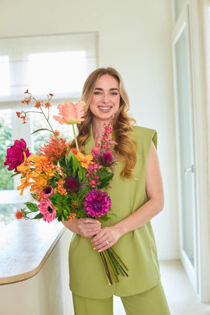 Une femme souriante aux longs cheveux blonds, vêtue d'une tenue vert clair, se tient à l'intérieur et tient le bouquet de fleurs artificielles en soie Magnus di Lambo — un bouquet coloré de fleurs en soie nouées à la main, parfait pour ceux qui préfèrent un arrangement floral sans entretien.