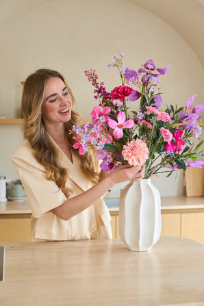 Une femme souriante aux longs cheveux blonds place un bouquet coloré de « Purple Dawn » composé de fleurs artificielles en soie dans un vase blanc sur le comptoir.