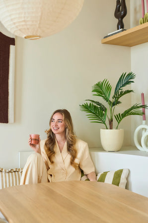Une femme souriante aux longs cheveux blonds est assise dans une pièce moderne et lumineuse avec une tasse rose à la main. Autour d'elle se trouvent des plantes vertes, dont la luxuriante plante artificielle Phoenix de 85 cm, ainsi que des étagères en bois et des objets décoratifs.