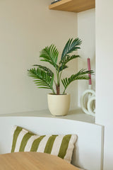 Une plante artificielle Phoenix de 85 cm en forme de palmier en pot se dresse sur une étagère blanche à côté d'une bougie rose et d'un objet de décoration blanc. En dessous, un coussin rayé vert et blanc repose sur un banc, tandis qu'une partie d'une table en bois est visible au premier plan.