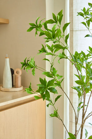 Une plante artificielle Pieris Japonica de 150 cm se dresse près d'une fenêtre à côté d'un meuble en bois clair contenant des bouteilles en céramique et un moulin à poivre, ajoutant une touche minimaliste à la pièce lumineuse et aérée.