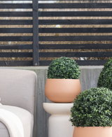 Trois topiaires rondes artificielles en buis de style romain UV D30 cm se trouvent sur un patio près d'une chaise d'extérieur beige, contre une clôture moderne à lattes noires et des arbres nus.