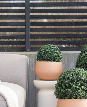 Trois topiaires rondes artificielles en buis de style romain UV D30 cm se trouvent sur un patio près d'une chaise d'extérieur beige, contre une clôture moderne à lattes noires et des arbres nus.