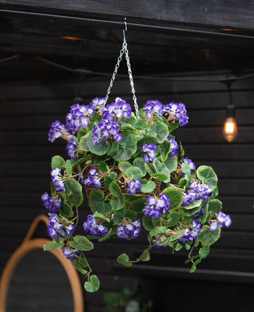 Le Bégonia artificiel violet en panier UV 70 cm est suspendu à une chaîne et présente des feuilles vertes luxuriantes et des fleurs violettes et blanches éclatantes. Derrière, un mur en bois sombre et une lampe lumineuse complètent la scène.